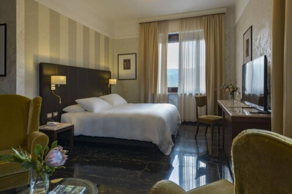 Ein Hotelzimmer mit einem großen Bett, einem Schreibtisch mit Fernseher, einem Sessel und einer Vase mit Blumen.