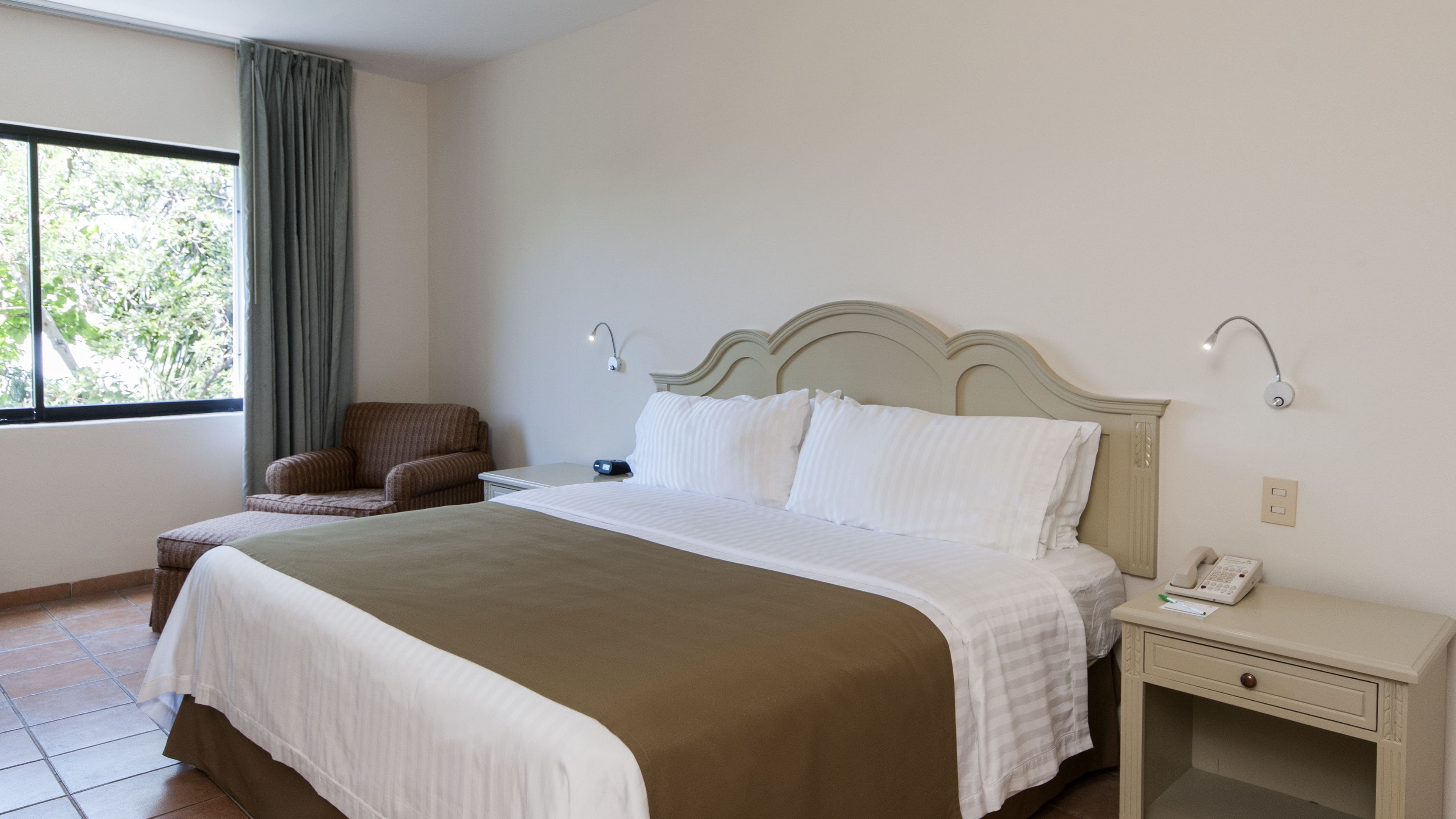 Quarto de hotel com uma cama grande, uma cadeira, mesa de cabeceira com telefone e janela com cortinas.