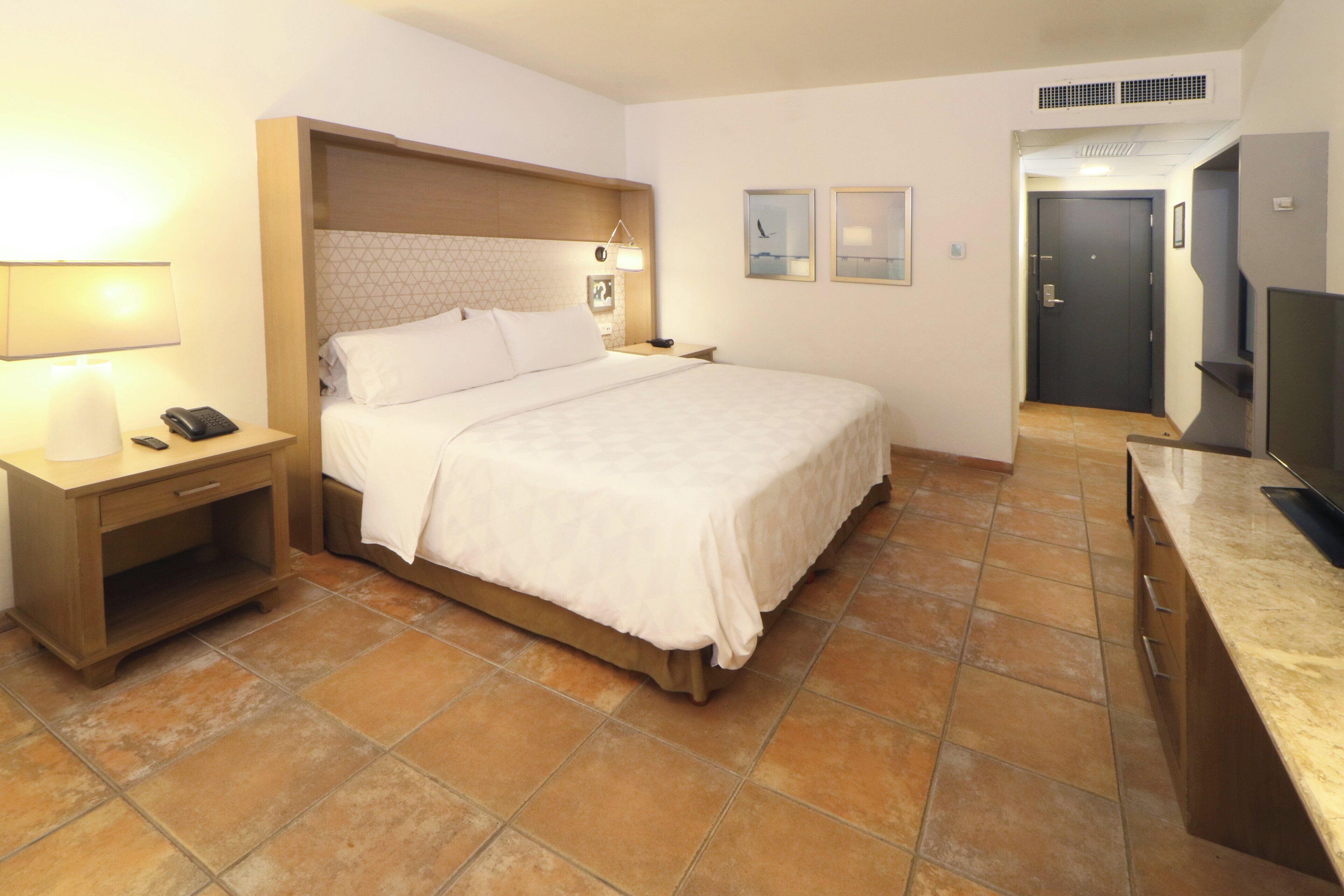 Habitación de hotel con cama, mesita de noche con lámpara, televisor y puerta que da a otra habitación.