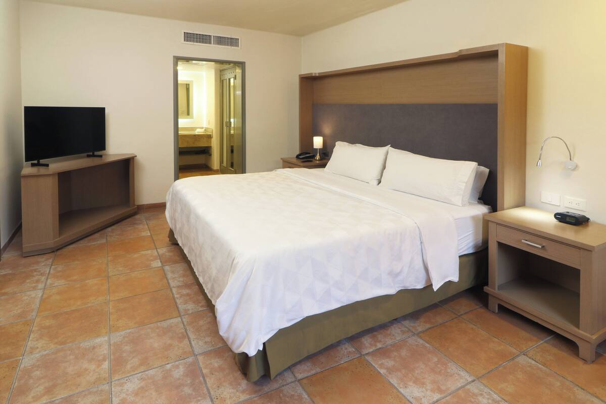 Una habitación de hotel con una cama grande, un televisor sobre un mueble bajo, una mesita de noche con lámpara y un baño visible a través de una puerta abierta.