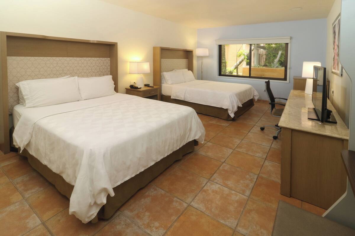 Habitación de hotel con dos camas, un escritorio con computadora y una ventana con vista a follaje.