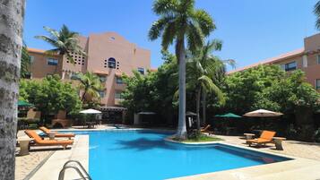 A swimming pool with lounge chairs and umbrellas, surrounded by palm trees and a building with a red-tiled roof.