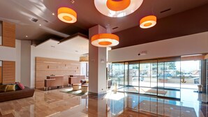 A modern hotel lobby with a wooden reception desk, a seating area with a sofa, and large windows offering a view of the street. - Hotel Vigo Los Galeones Affiliated by Meliá (Vigo)
