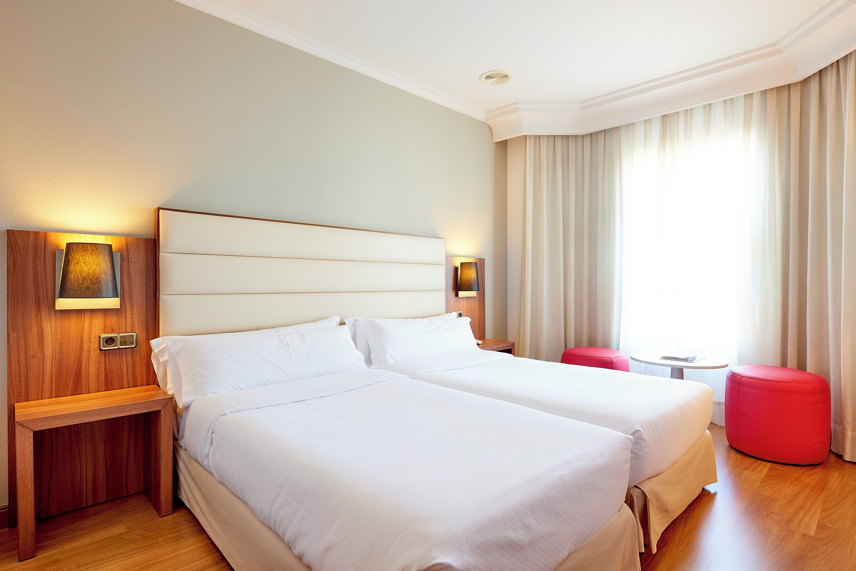 Habitación de hotel con dos camas, una mesita de noche de madera, un reposapiés rojo y una pequeña mesa con una lámpara.