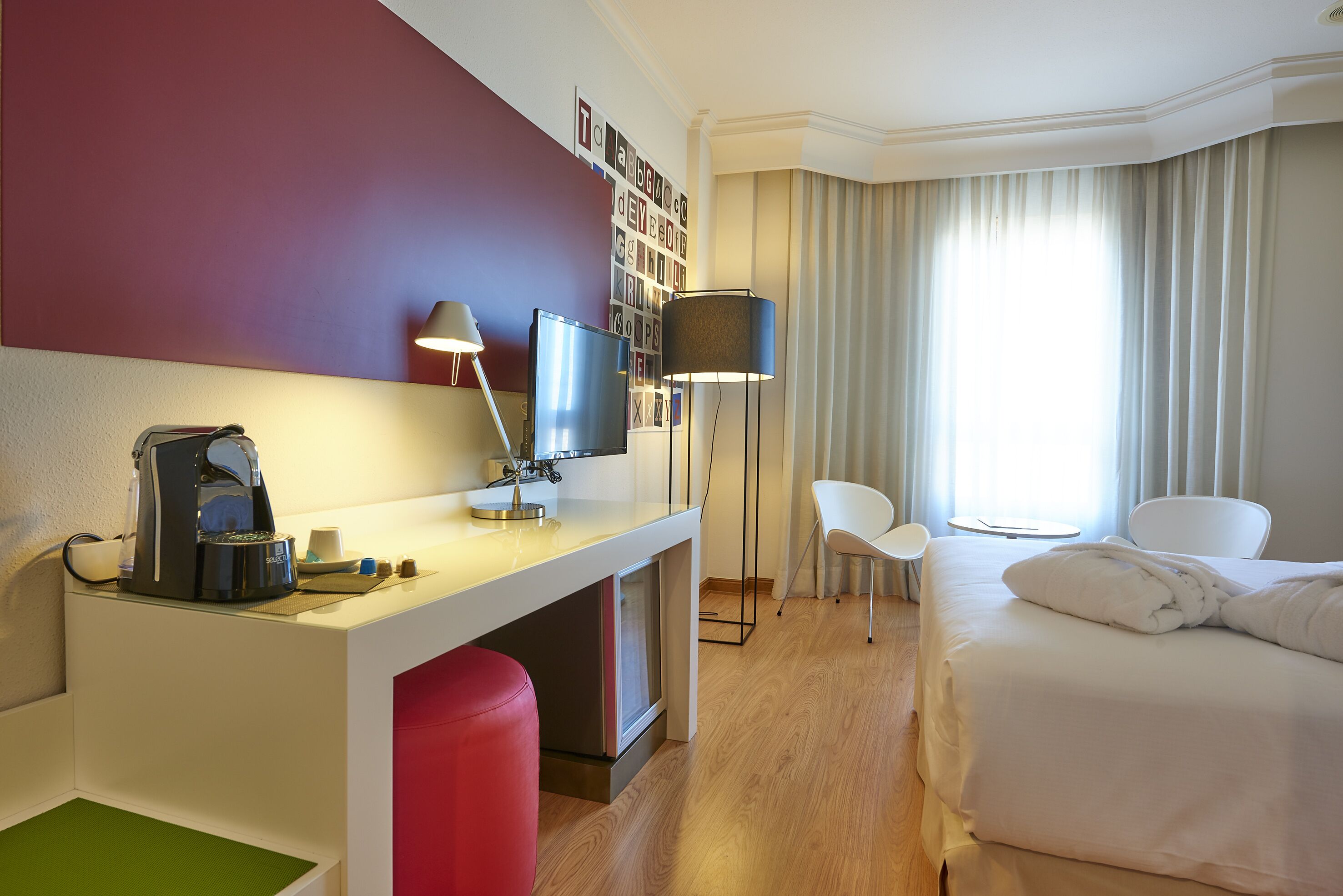 Habitación de hotel con cama, escritorio con cafetera, televisor, silla y ventana con cortinas.