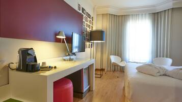 Habitación de hotel con cama, escritorio con cafetera, televisor, silla y ventana con cortinas.