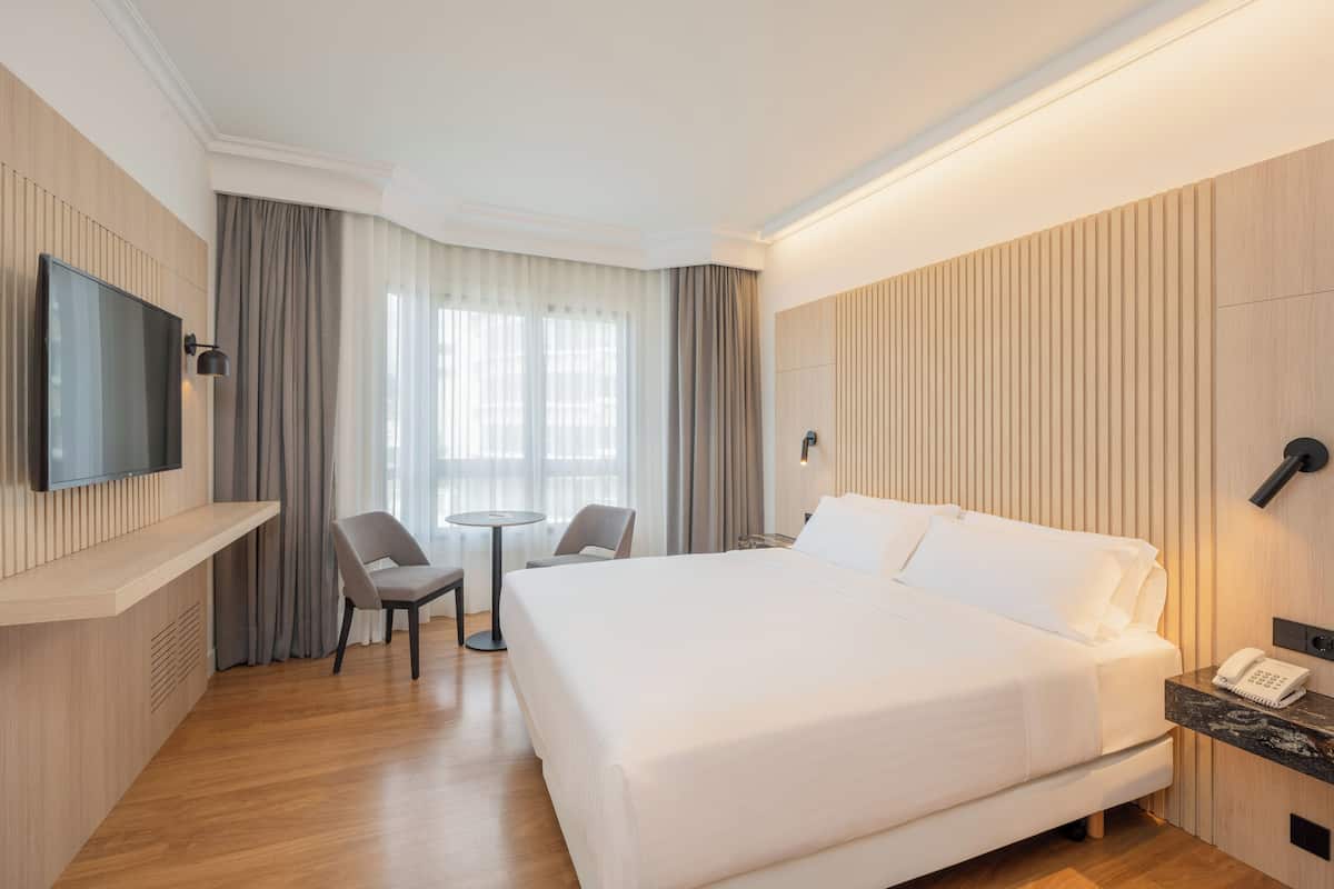 A hotel room with a large bed, a flat-screen TV mounted on the wall, a small round table, two chairs, a desk with a telephone, and a window with curtains.