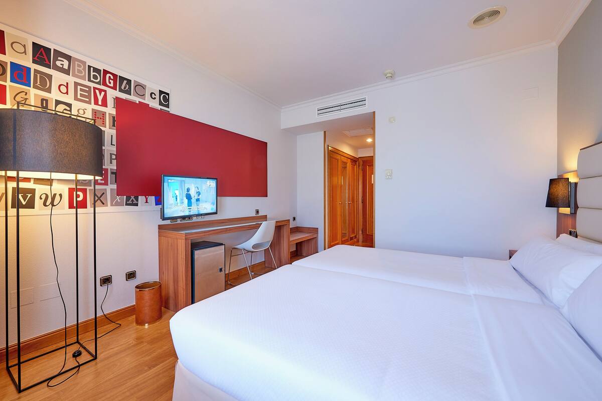 A hotel room with a bed, desk, chair, TV, and a wall with alphabet letters.