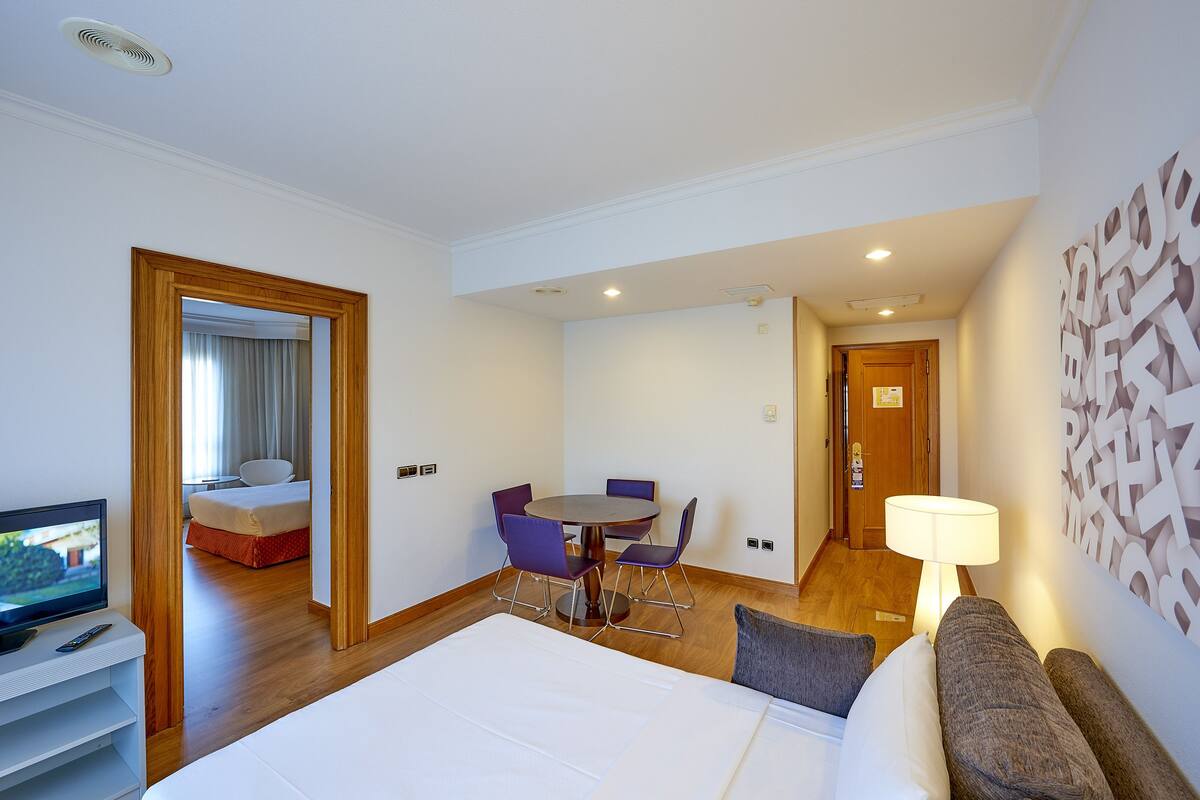 A hotel room with a bed, a TV, a dining table with chairs, and a lamp.