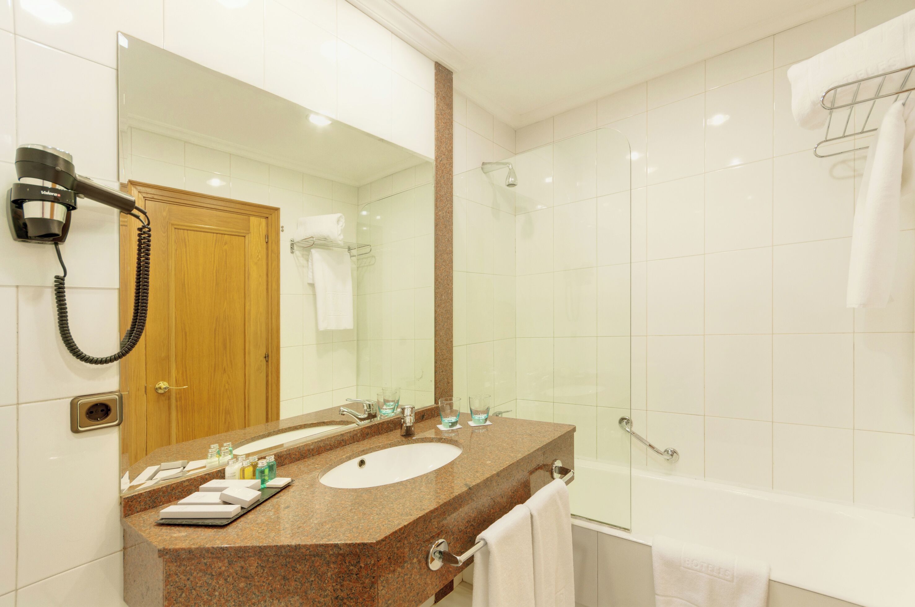 Casa de banho com lavatório, espelho e chuveiro. Um secador de cabelo está fixado na parede e há um porta-toalhas.