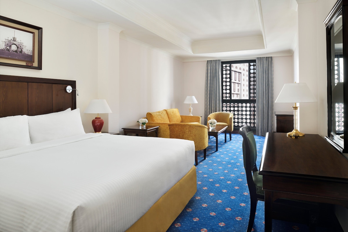 Ein Hotelzimmer mit einem großen Bett, zwei Sesseln, einem Schreibtisch und einem Fenster mit Blick auf ein Gebäude.