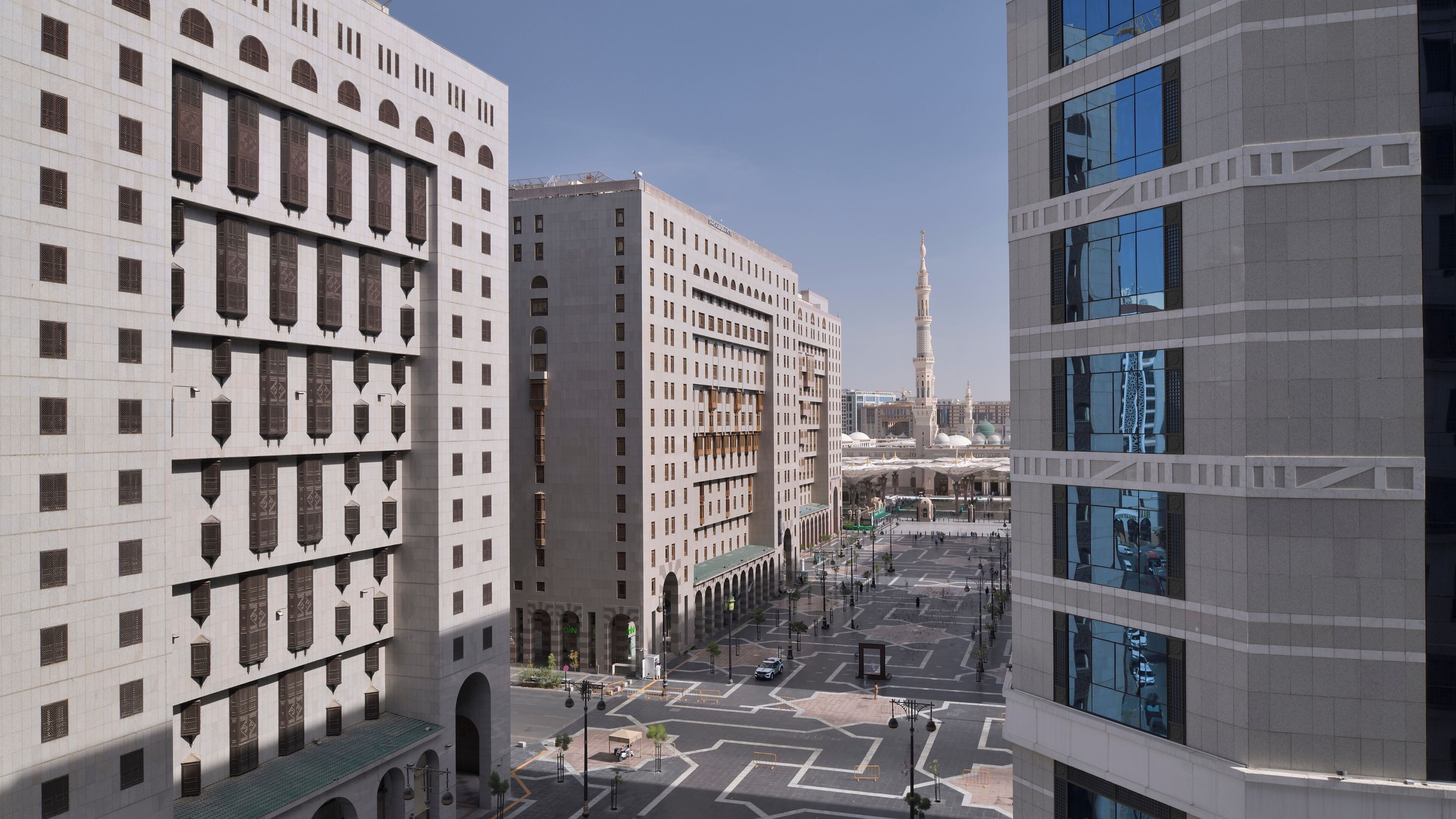 A modern urban landscape with a prominent mosque and minaret in the background, surrounded by contemporary buildings with large windows and a clear sky.