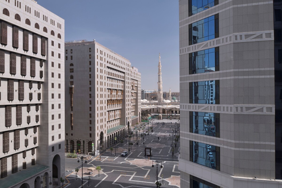 Een modern stadslandschap met een opvallende moskee en minaret op de achtergrond, omringd door moderne gebouwen met grote ramen en een heldere lucht.