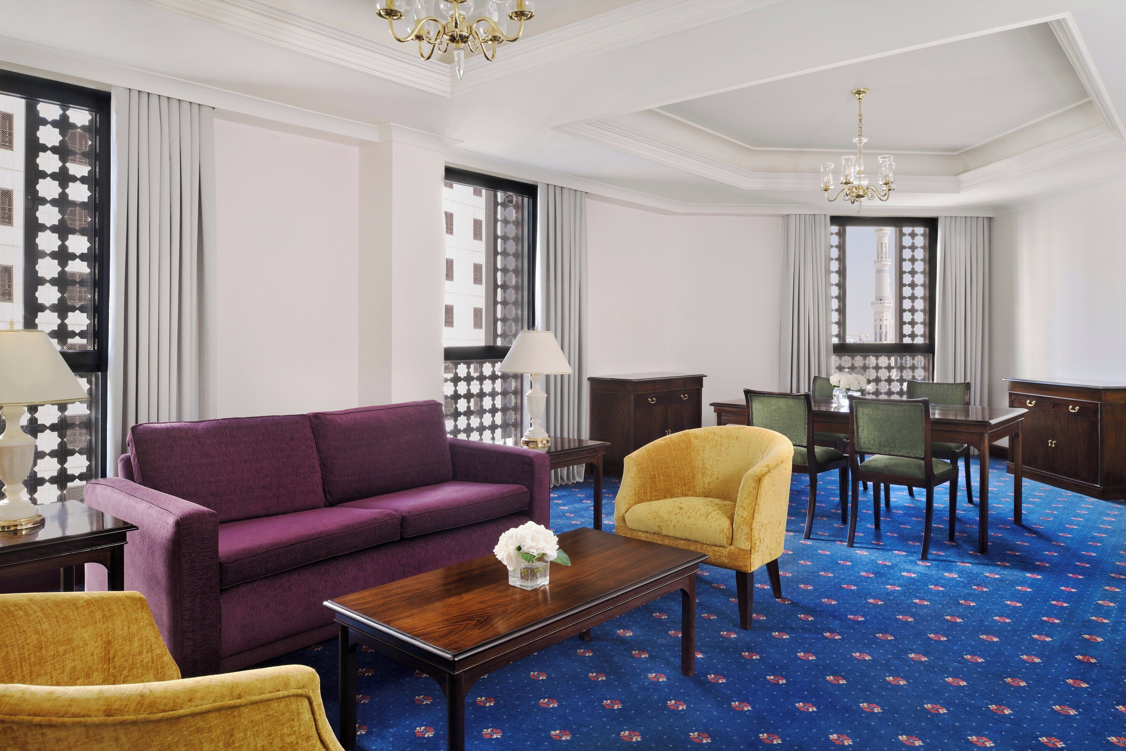 A hotel room with a purple sofa, yellow armchair, and a dining area with a table and chairs.