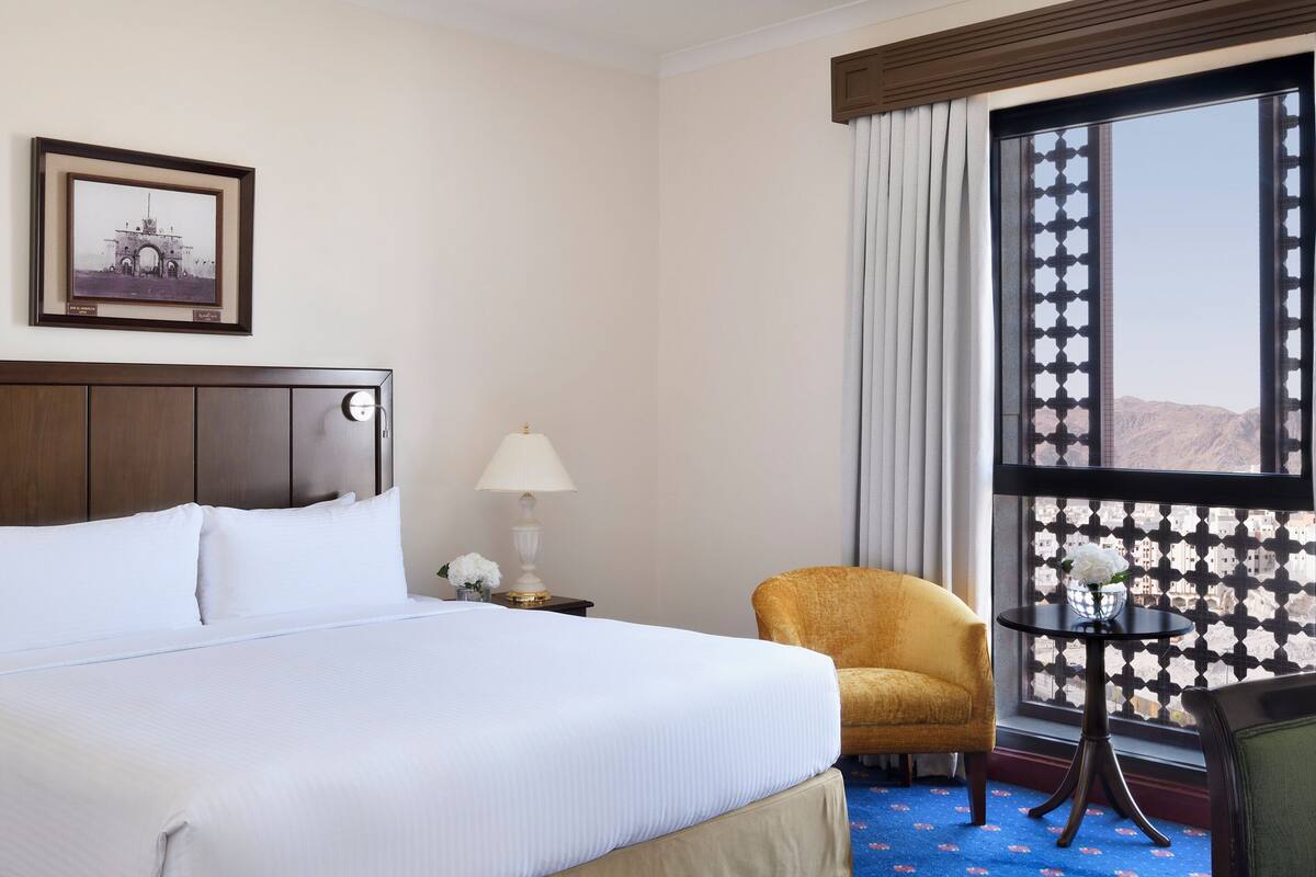 Une chambre d’hôtel avec un grand lit, une chaise, une petite table, une lampe, une fenêtre donnant sur un beau paysage et un tableau encadré au mur.