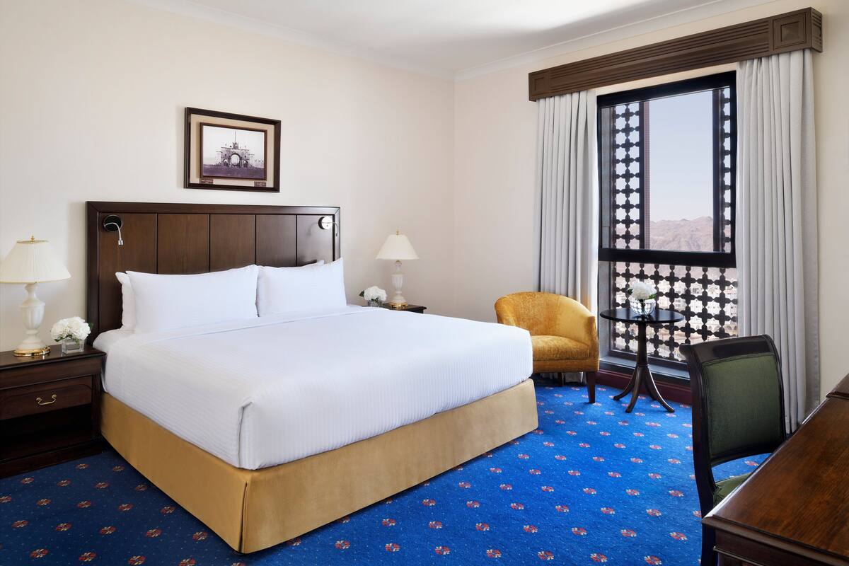 Une chambre d’hôtel avec un grand lit, une chaise, un bureau, une fenêtre avec des rideaux et un tableau encadré au mur.