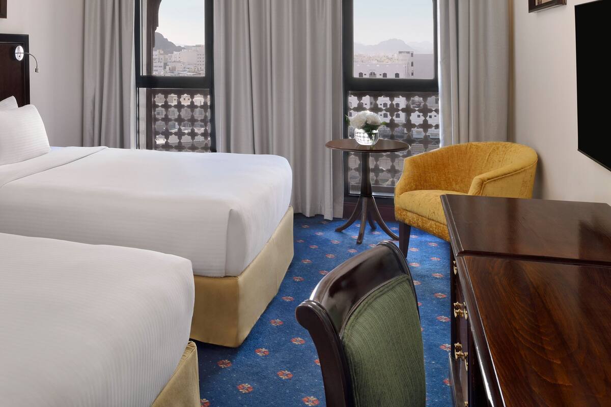 Une chambre d’hôtel avec deux lits, une petite table sur laquelle est posé un vase de fleurs, un fauteuil jaune et un téléviseur fixé au mur.