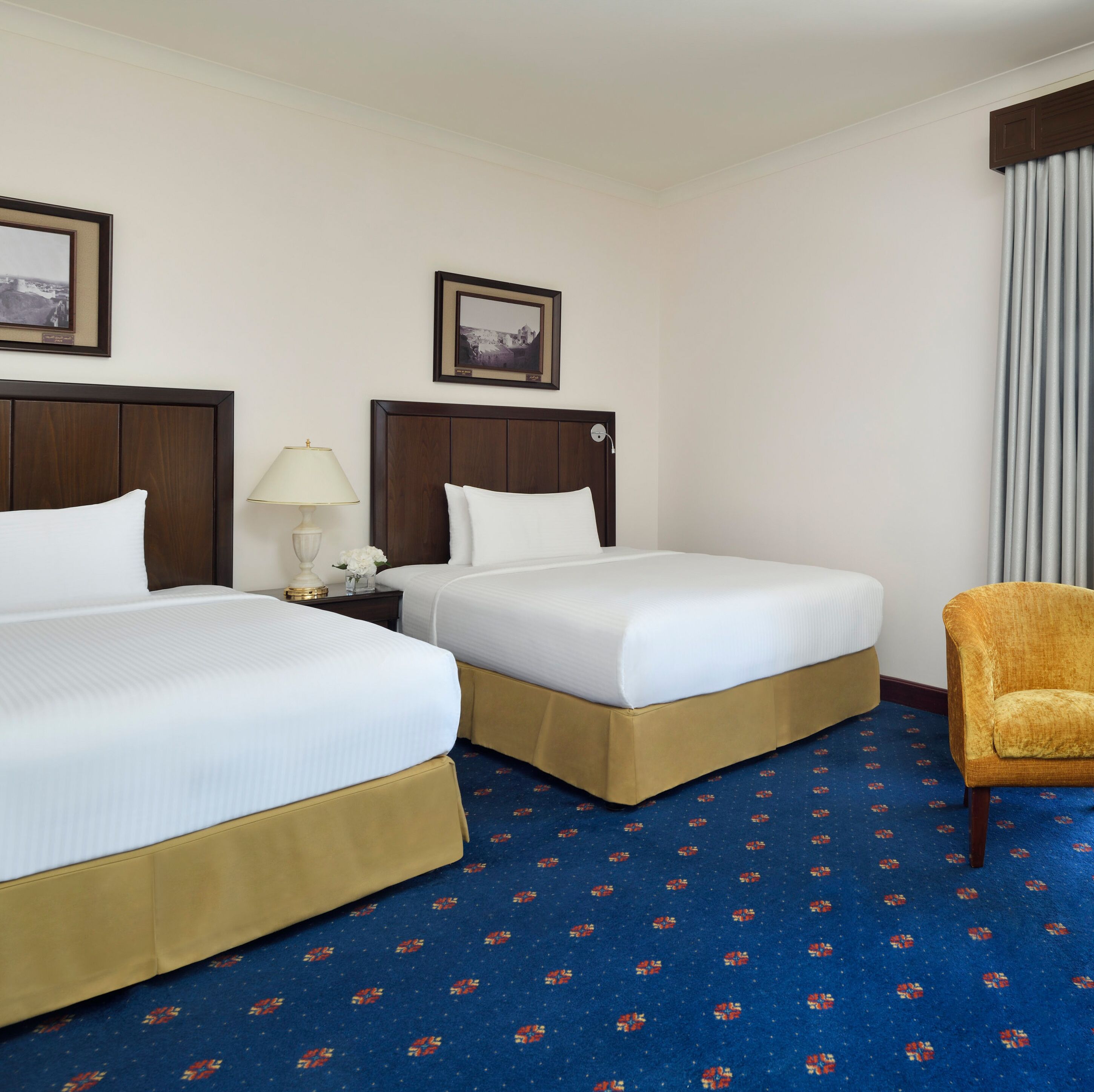 Een hotelkamer met twee bedden, een nachtkastje met een lamp, een stoel en een kamerbreed tapijt met een patroon.