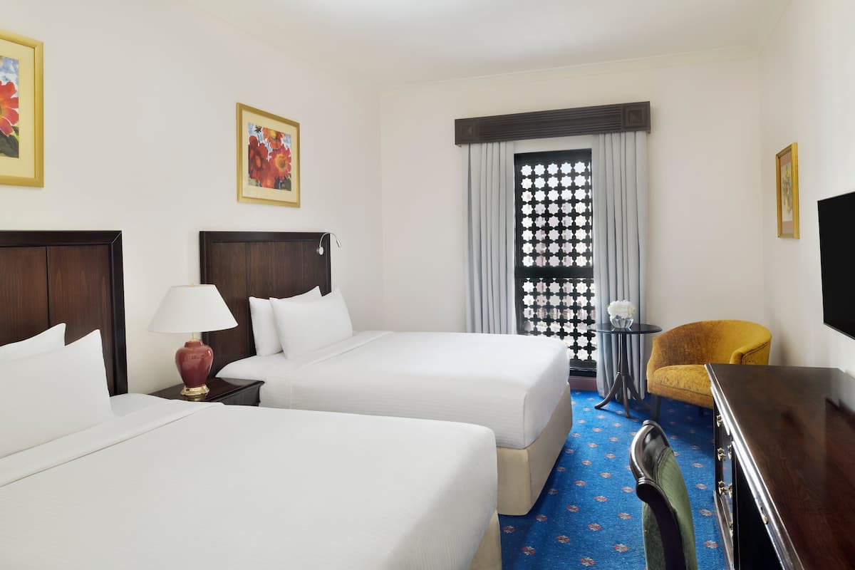 Une chambre d’hôtel avec deux lits, un bureau, une chaise, une lampe et une fenêtre avec des rideaux.