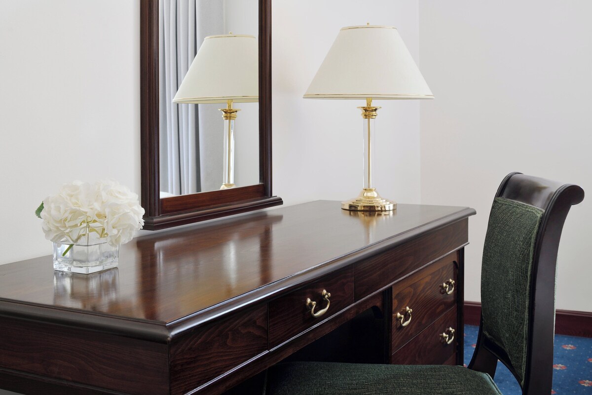 Een houten bureau met een spiegel, twee lampen, een vaas met witte bloemen en een groene fauteuil.