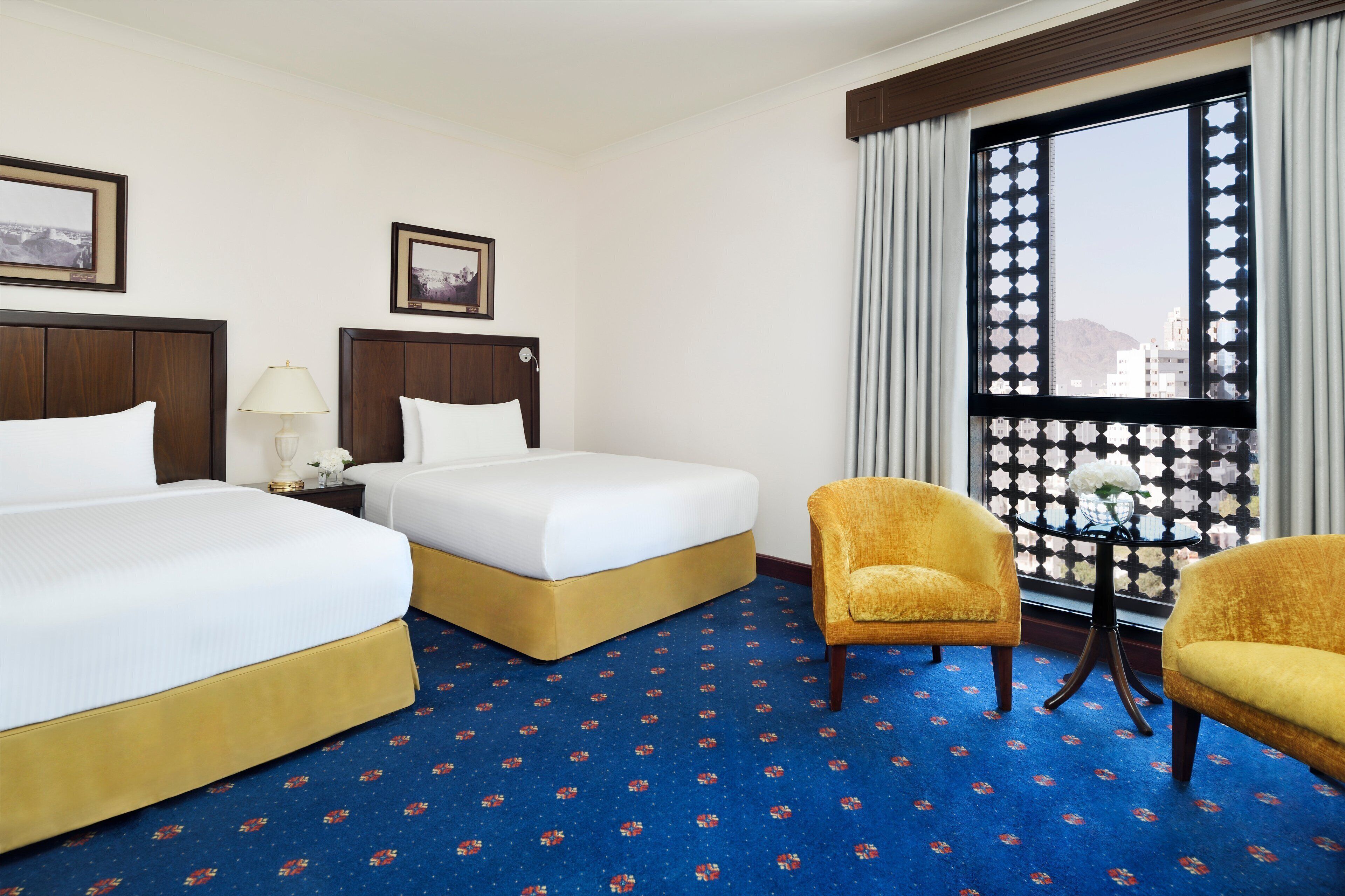 A hotel room with two beds, a small table with a lamp, a chair, and a window with a view of buildings.