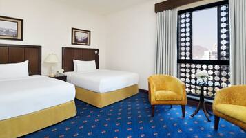 A hotel room with two beds, a small table with a lamp, a chair, and a window with a view of buildings.