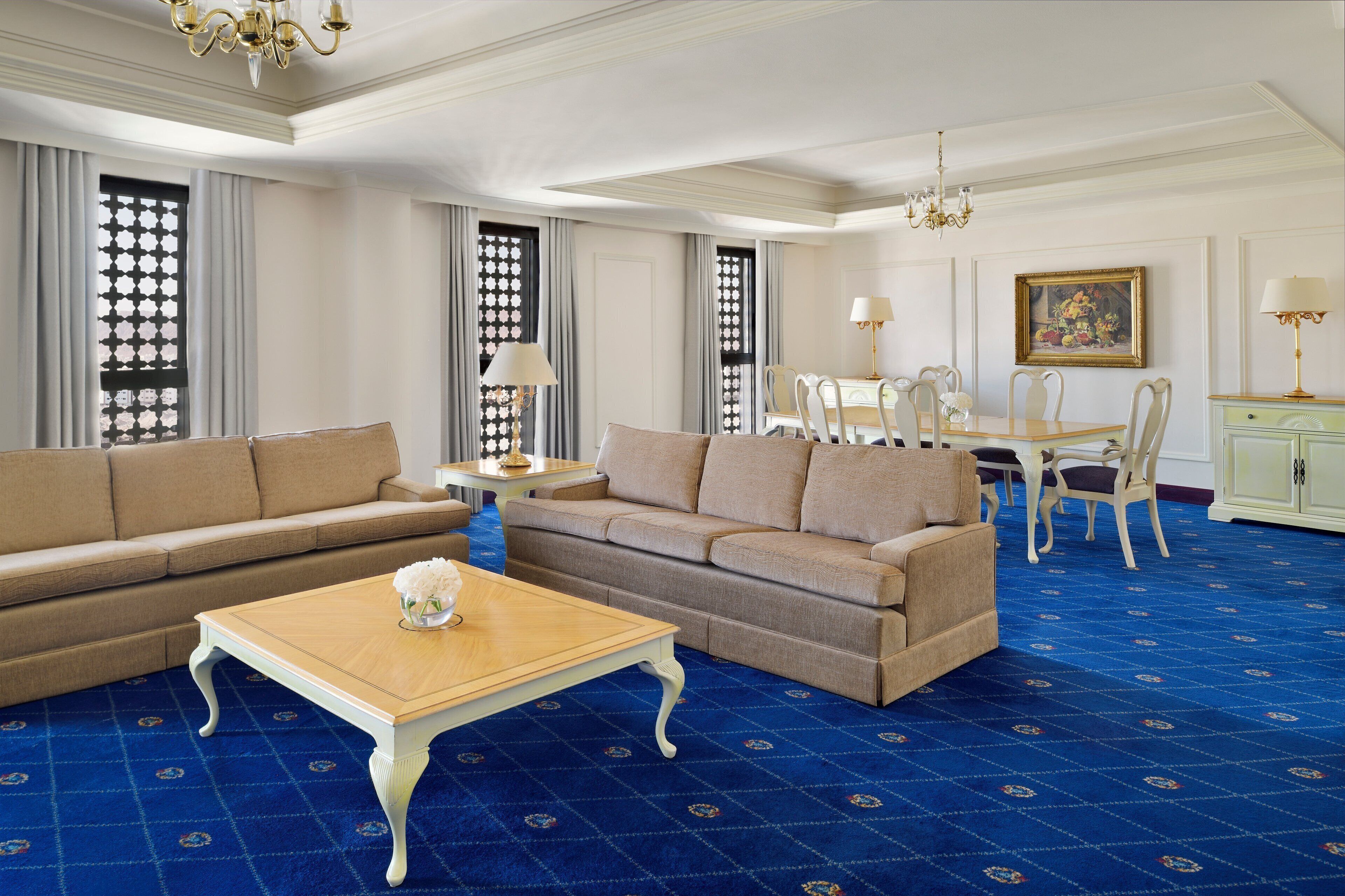 A spacious living room with a blue patterned carpet, beige sofas, a wooden coffee table, and a dining area with a white table and chairs.