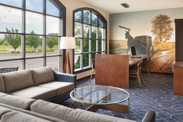 A modern hotel room with a large window, a sofa, a glass coffee table, a desk, and a mural of a horse-drawn carriage. - Courtyard by Marriott Waterloo St. Jacobs (Township of Woolwich)