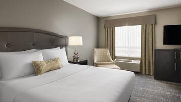 A hotel room with a large bed, a bedside lamp, a chair, a television, and an air conditioning unit.