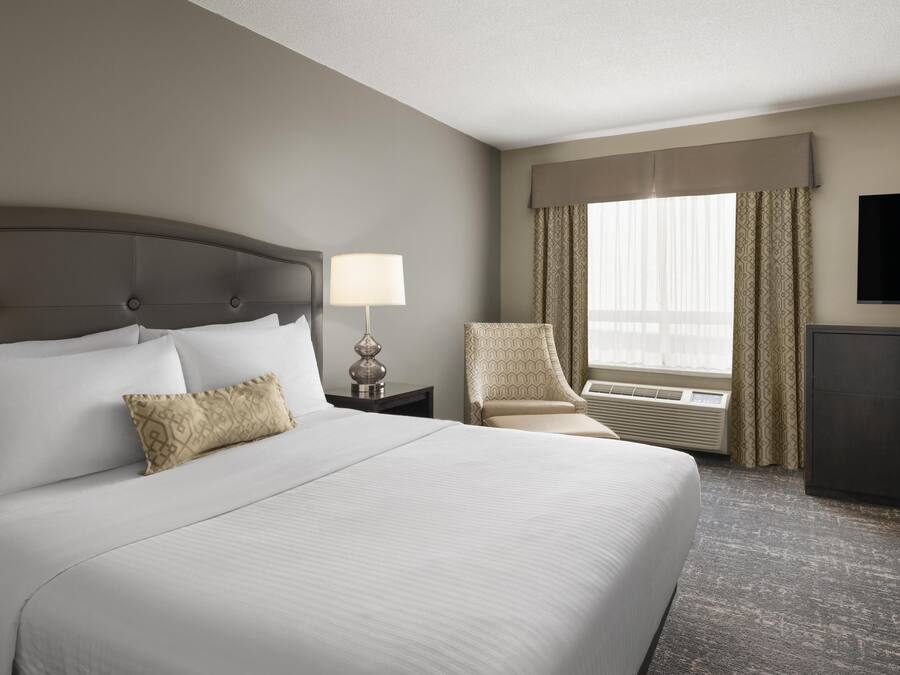 A hotel room with a large bed, a bedside lamp, a chair, a television, and an air conditioning unit.