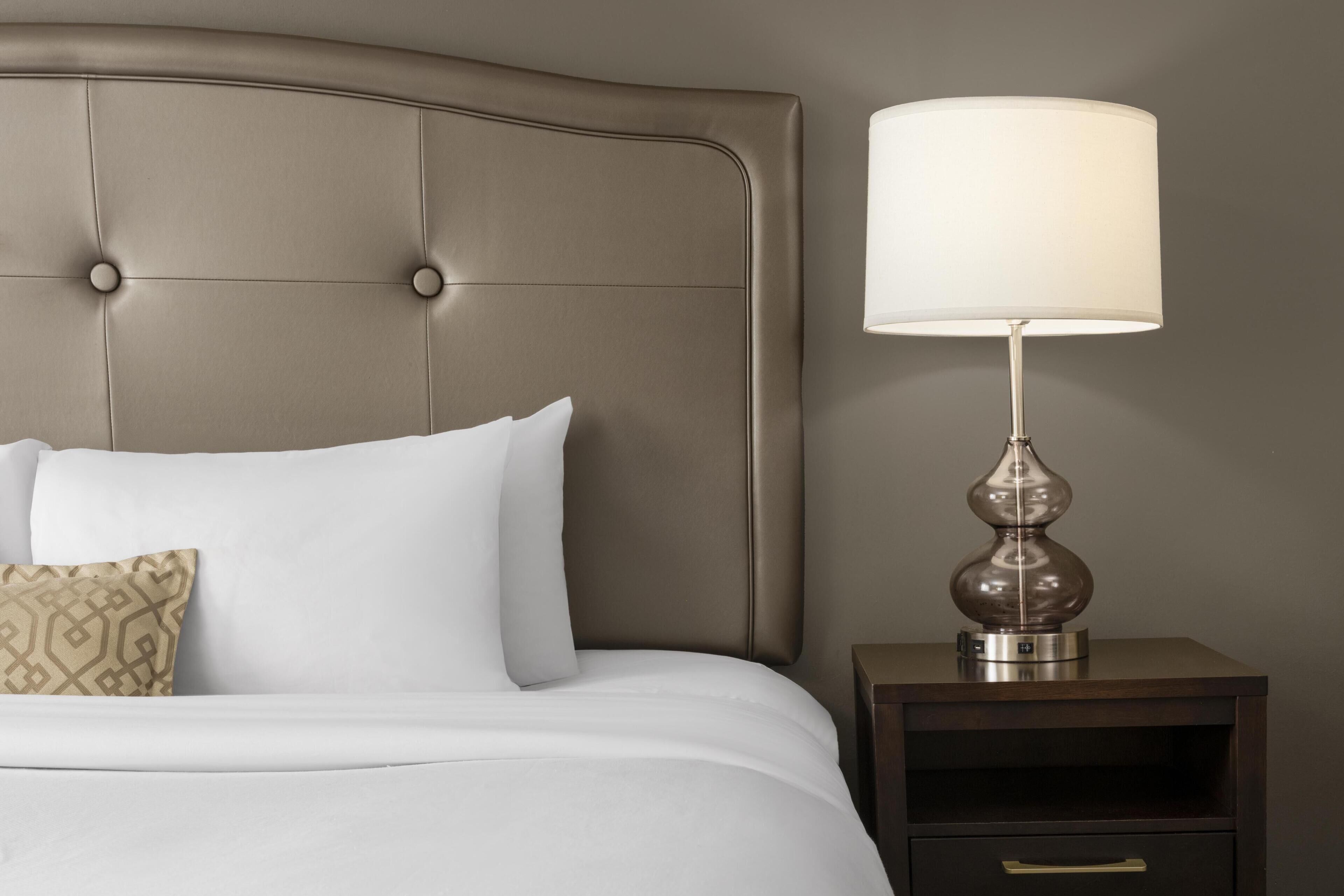 Un lit avec une literie blanche et un seul coussin à motifs, une lampe de chevet et une table de chevet en bois foncé avec un tiroir.