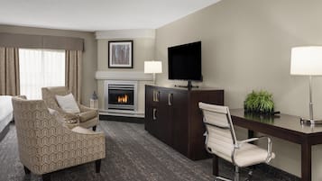 Une chambre d’hôtel comprenant un lit, un fauteuil à motifs, un bureau avec une chaise, un téléviseur à écran plat, une cheminée et une plante.