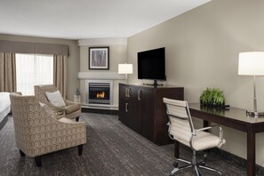 A hotel room with a bed, a patterned armchair, a desk with a chair, a flat-screen TV, a fireplace, and a plant. - Best Western Swan Castle Inn (Cochrane)