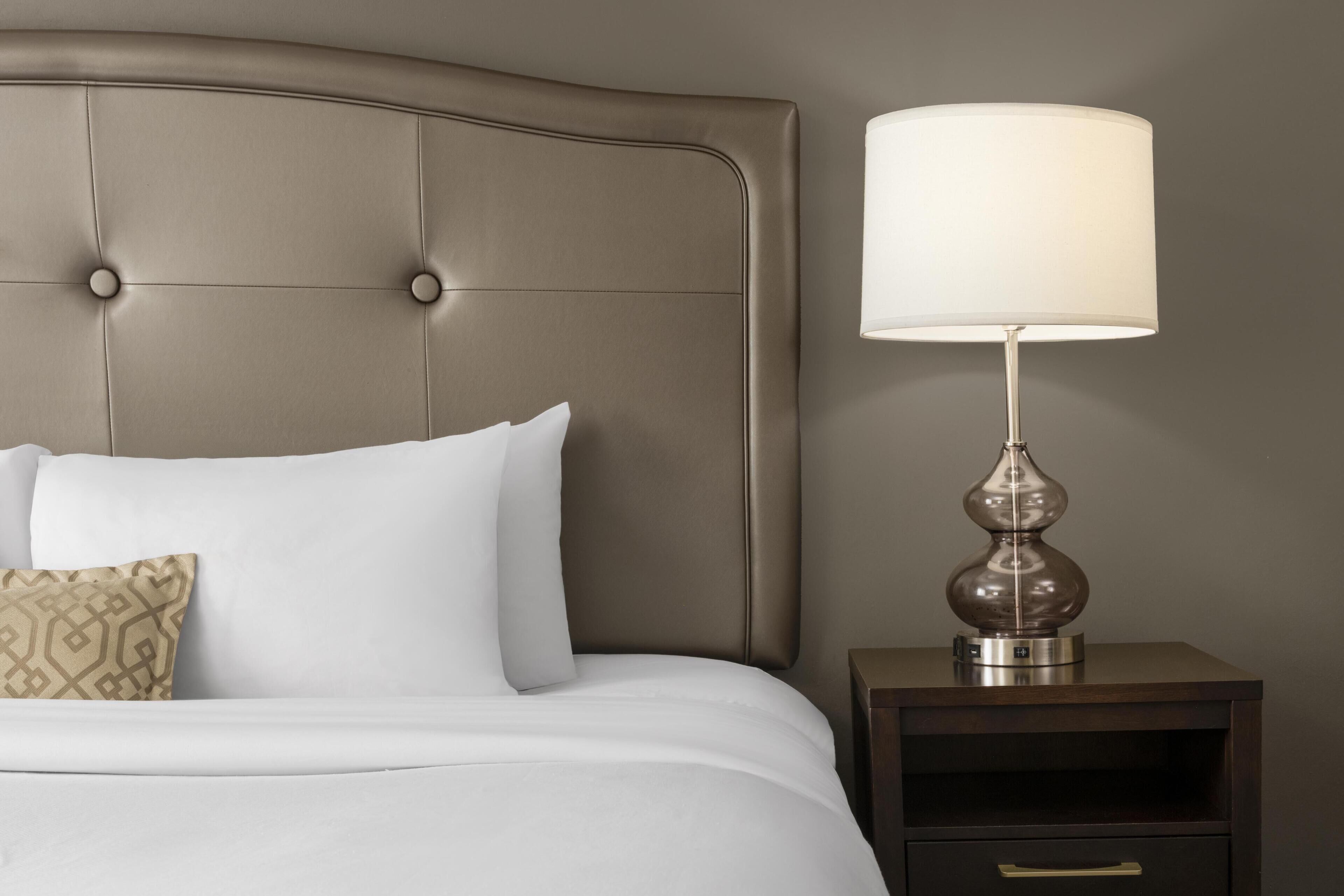 A neatly made bed with white linens and a single patterned pillow, a bedside lamp with a clear shade, and a small wooden nightstand with a drawer.