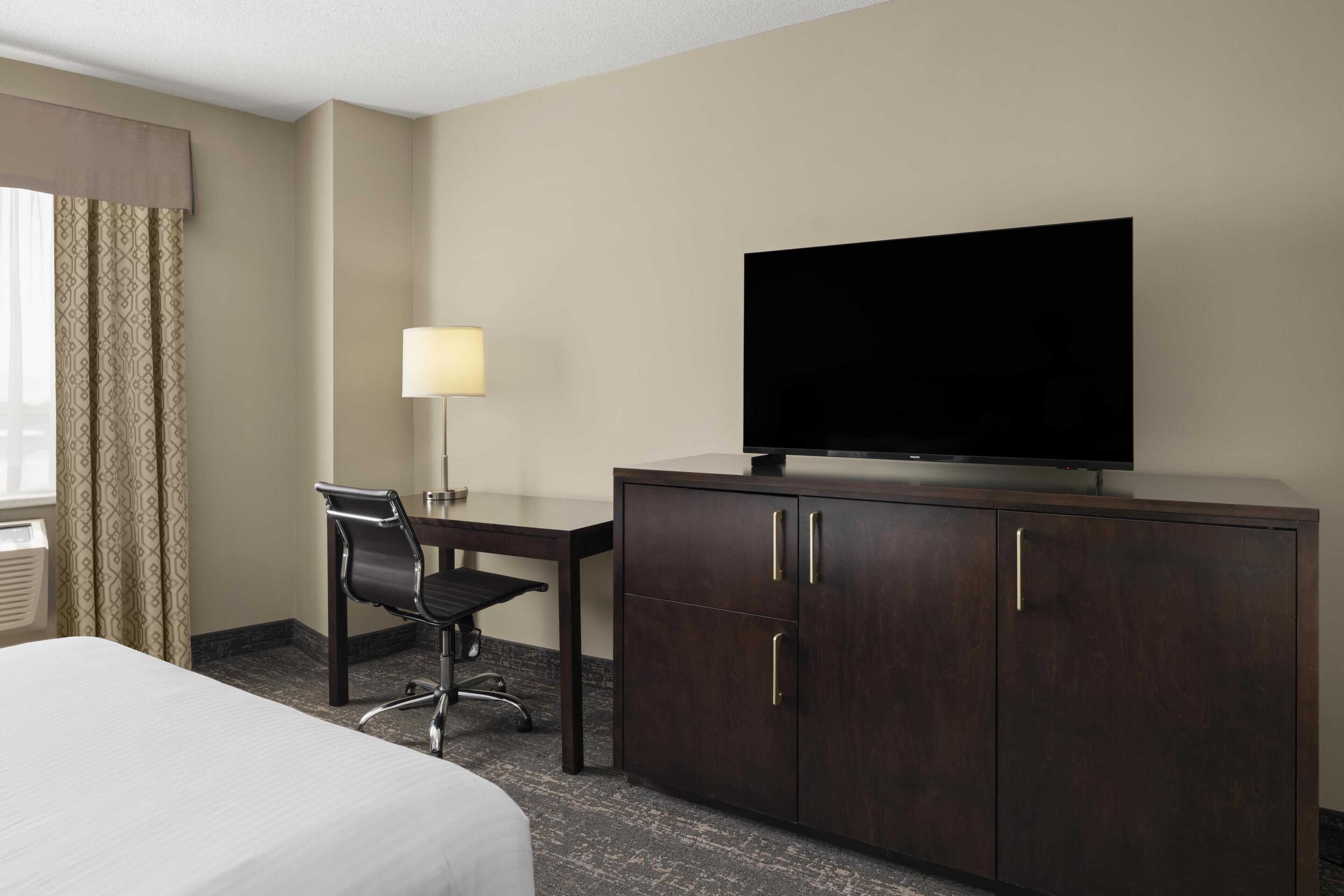 A hotel room with a bed, a desk with a chair, a television on a cabinet, and a lamp.