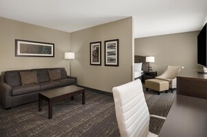 Habitación de hotel con sofá, sillón, reposapiés y escritorio. Se observan dos cuadros en la pared y una lámpara sobre el escritorio.