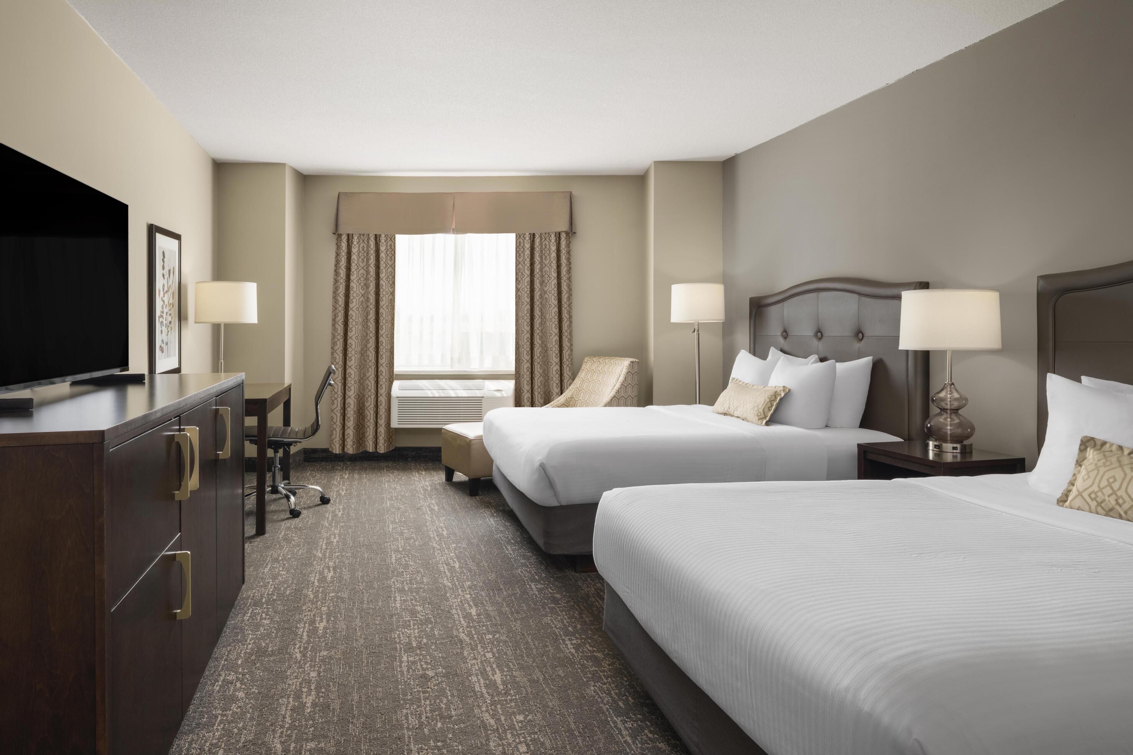 A hotel room with two beds, a desk, a television, and a window with curtains.
