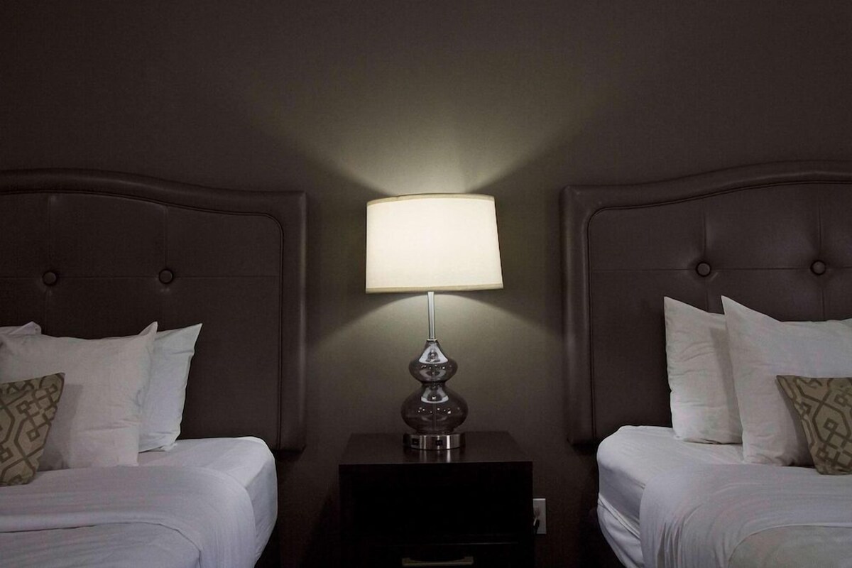 A hotel room with two beds, a nightstand with a lamp, and a wall-mounted light fixture.