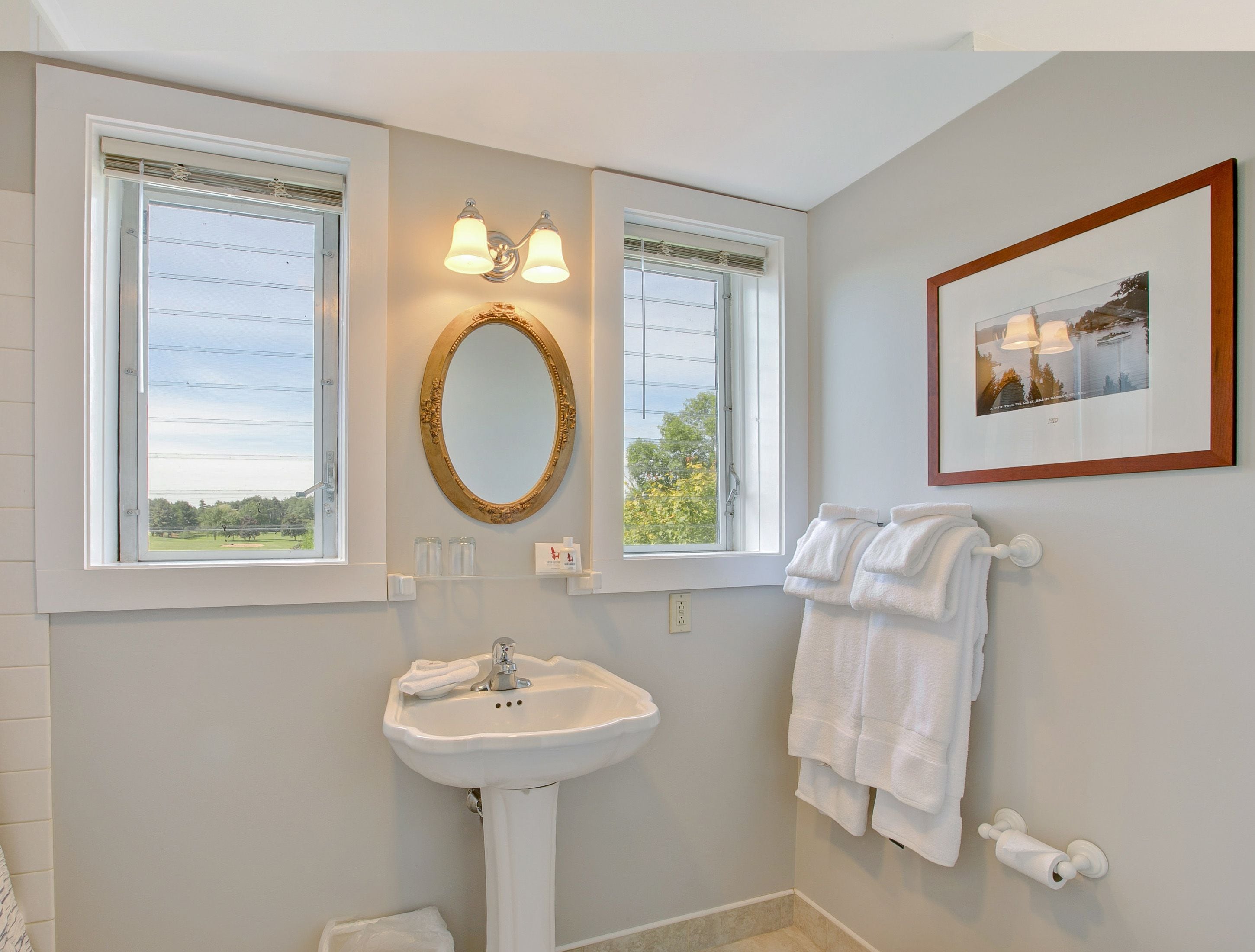 A bathroom with a pedestal sink, two windows with white frames, a wall-mounted mirror with a gold frame, and a framed picture on the wall.