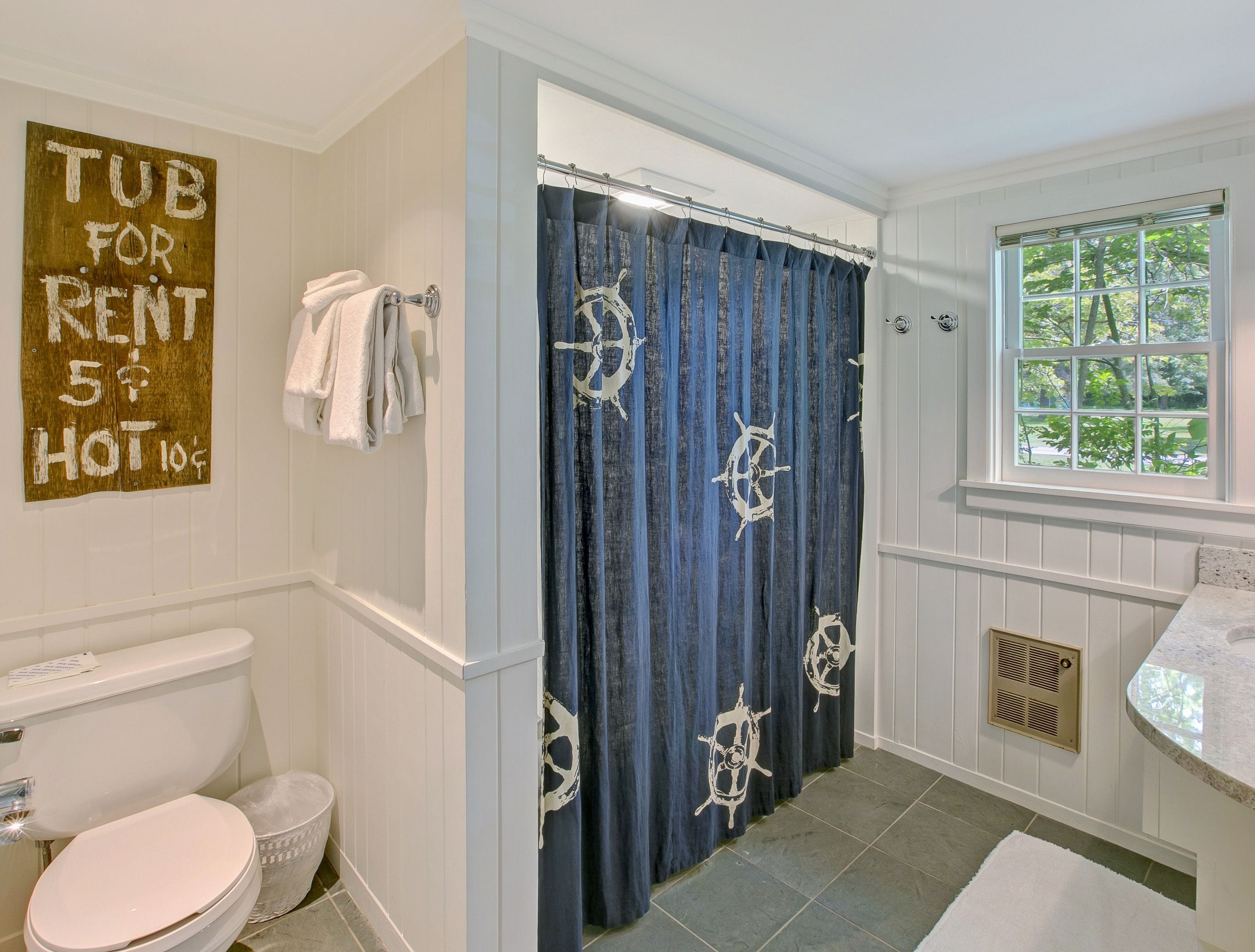 A bathroom with a toilet, a shower curtain with a nautical theme, a window with a view of trees, and a sign on the wall.