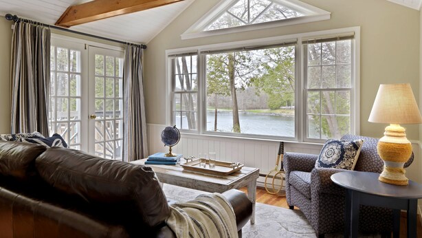Una sala de estar acogedora con un sofá de cuero, un sillón estampado, una mesita con lámpara y ventanas amplias que dan a vista de árboles y un cuerpo de agua.