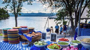 Una mesa de buffet con diversos alimentos y platos coloridos, ubicada cerca de un lago con una carpa al fondo.