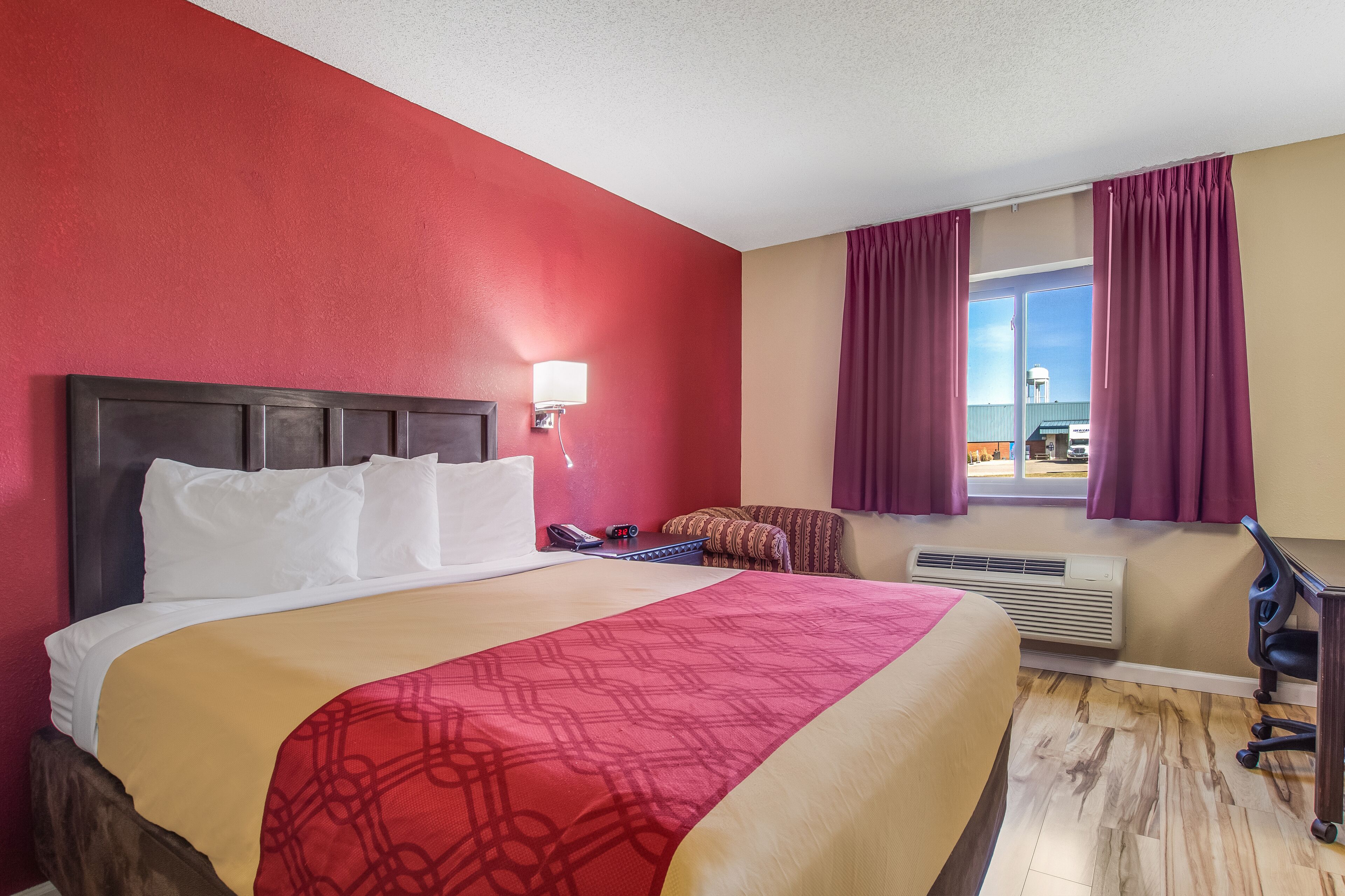 A hotel room with a large bed, a desk, a chair, a window with curtains, a wall-mounted lamp, and a view of a water tower outside.