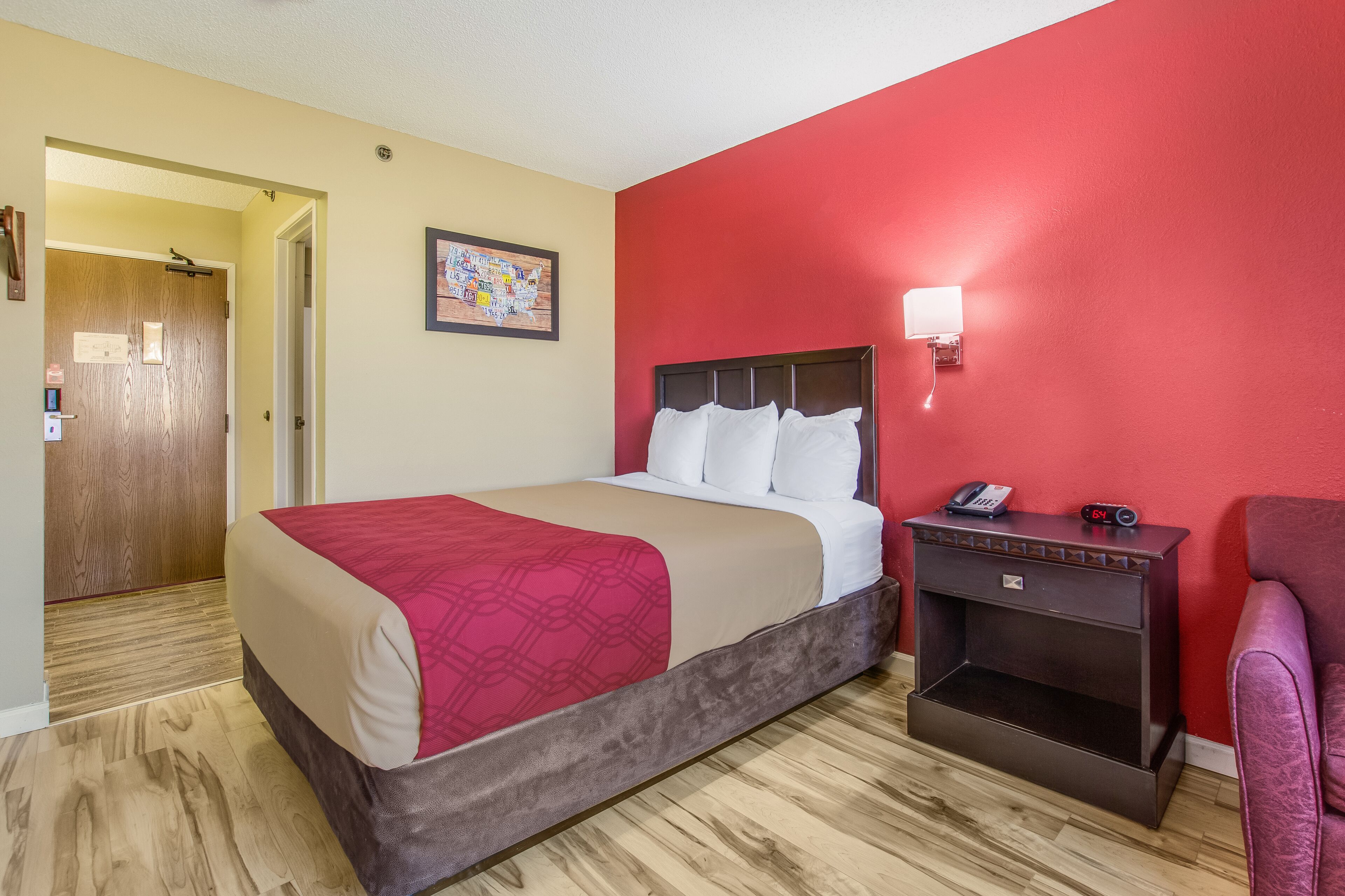 A hotel room with a bed, a nightstand, a chair, a red wall, and a wooden door.