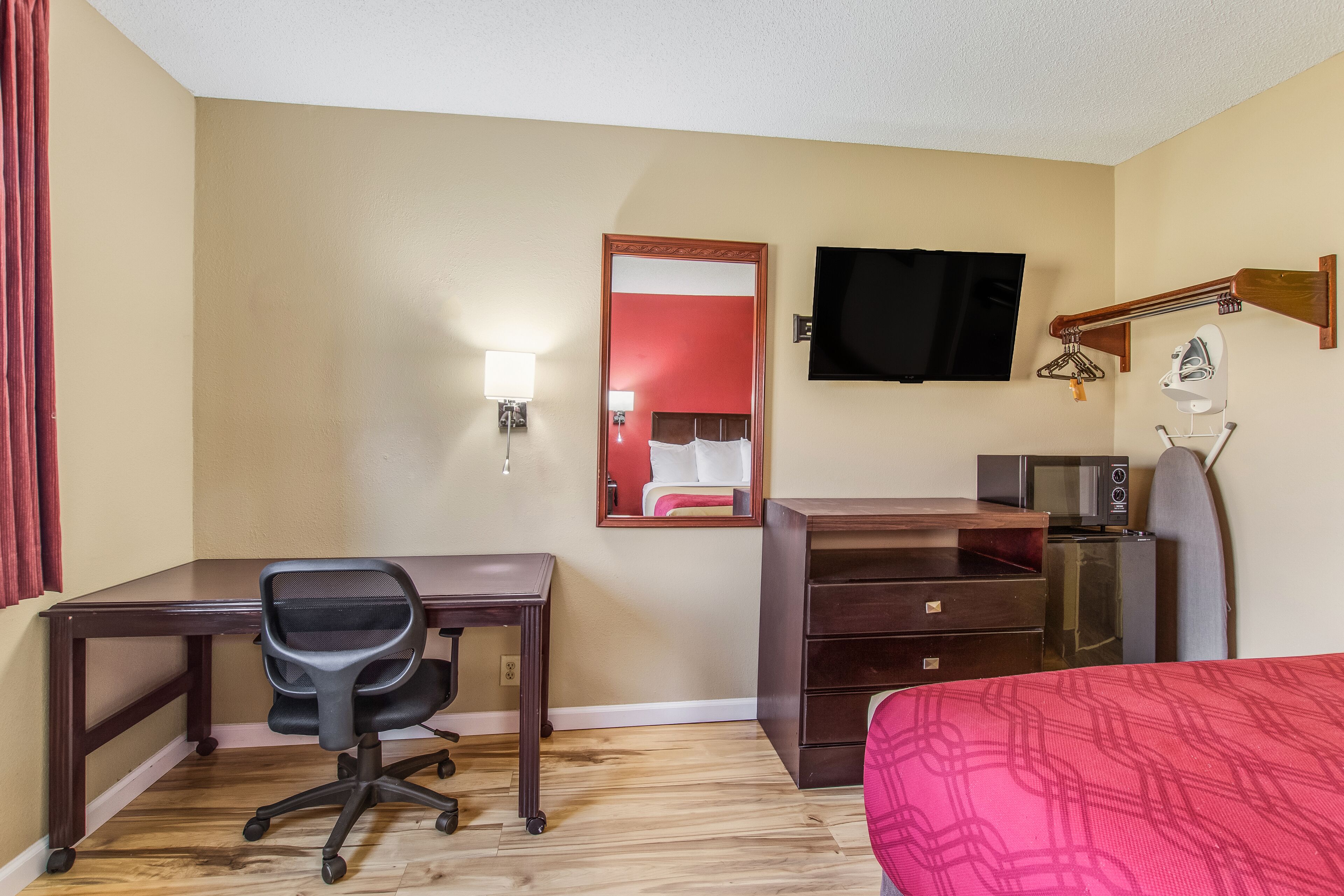 A hotel room with a wooden desk, a black office chair, a bed with a red patterned bedspread, a television mounted on the wall, a microwave, and a small dresser.