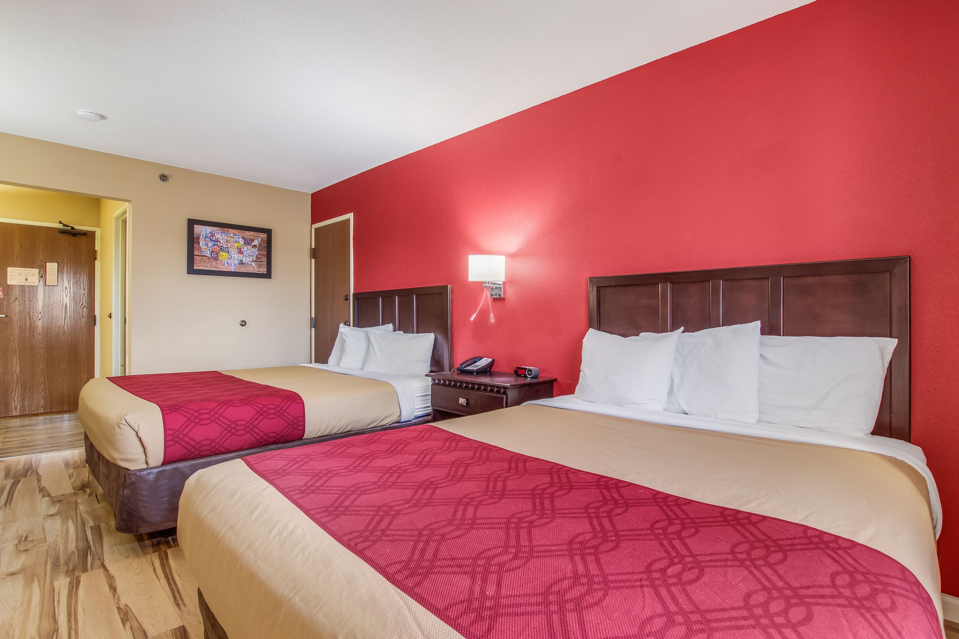 A hotel room with two beds, a red wall, a wooden door, and a nightstand with a phone and a cup.