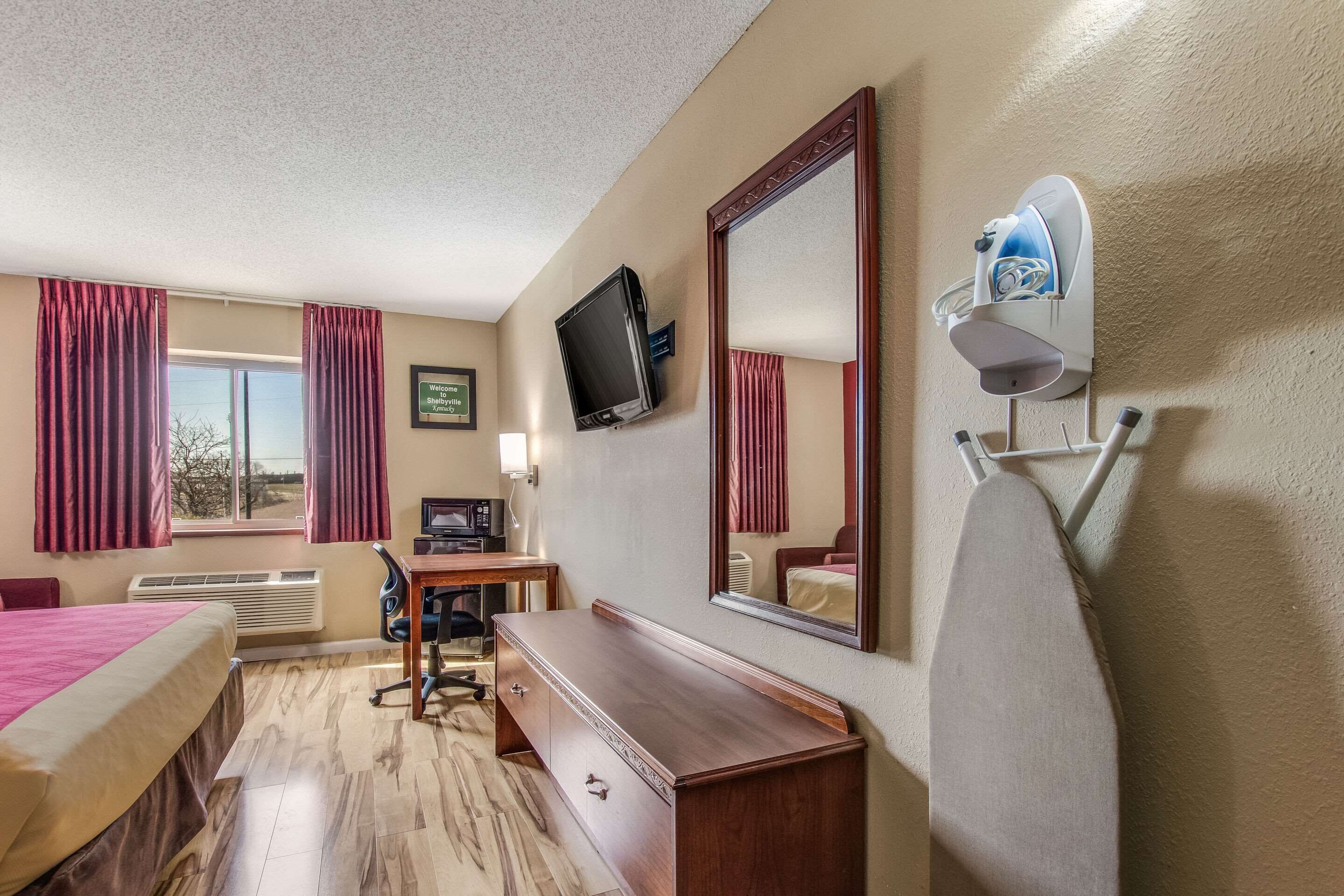 A hotel room with a bed, desk, chair, TV, mirror, and a hairdryer on the wall.