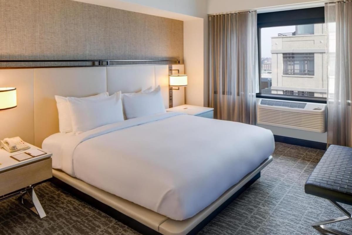 Una habitación de hotel con una cama grande, mesitas de noche, un escritorio, una silla, una ventana con cortinas y un equipo de aire acondicionado.