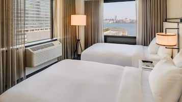 A hotel room with two beds, a large window with a city view, a standing lamp, and a heater.
