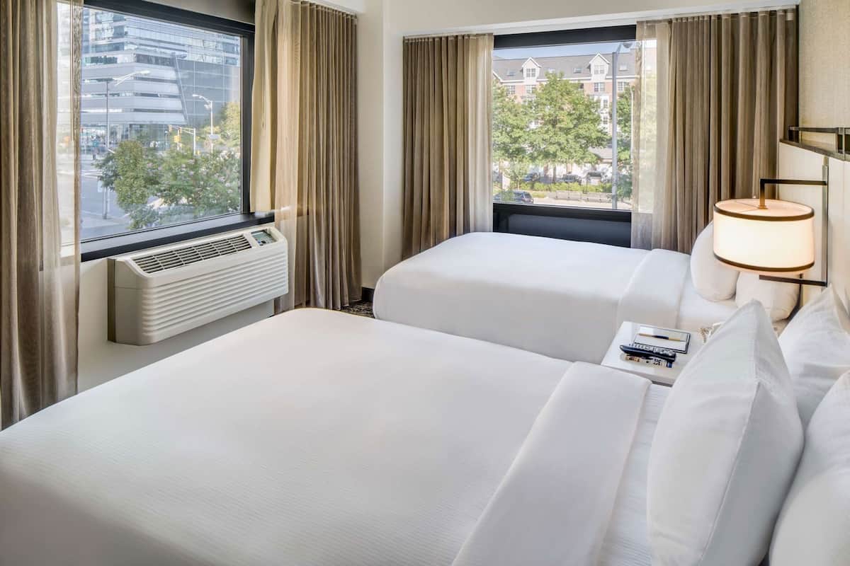 A hotel room with two beds, a window with curtains, an air conditioning unit, and a bedside lamp.